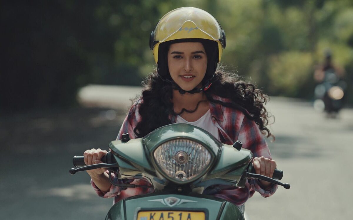 A frame from a Denzong Films brand film: a young woman riding a scooter along a tree lined road