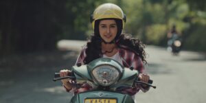 A frame from a Denzong Films brand film: a young woman riding a scooter along a tree lined road